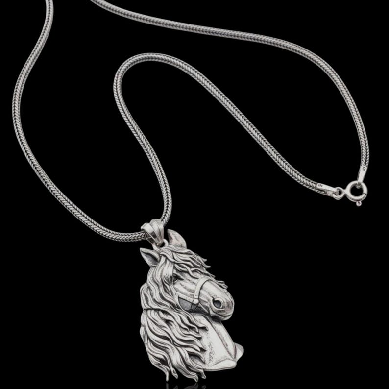 Puede incluir: Un collar plateado con un colgante detallado de cabeza de caballo. El colgante muestra el perfil del caballo con una melena y rasgos faciales detallados. La cadena tiene un dise&ntilde;o tejido y el cierre es visible. El fondo es negro.