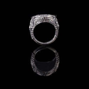 Op de afbeelding: Een zilveren ring met ingewikkelde ontwerpen en een verhoogd midden. De ring ligt op een zwarte achtergrond en reflecteert licht.