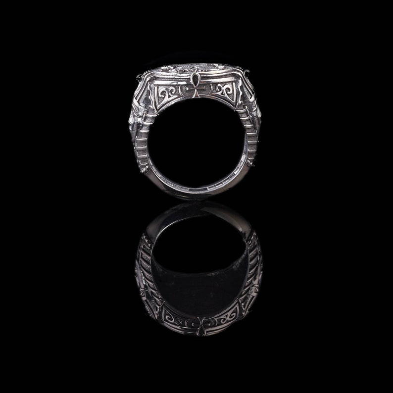 Op de afbeelding: Een zilveren ring met ingewikkelde ontwerpen en een verhoogd midden. De ring ligt op een zwarte achtergrond en reflecteert licht.