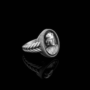 May include: A silver ring with a braided band and a round, raised cameo featuring a person wearing a cap with a star.