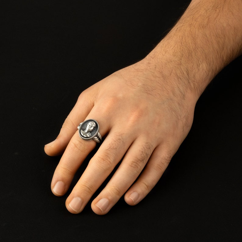 May include: A silver ring with a detailed oval-shaped design featuring a face in relief. The ring is worn on a person's finger.