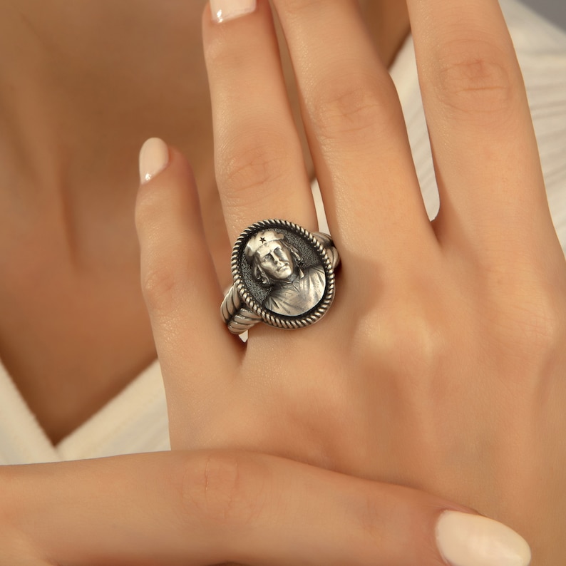 May include: A silver ring with a detailed oval design featuring a portrait of a person with a cross on their head.