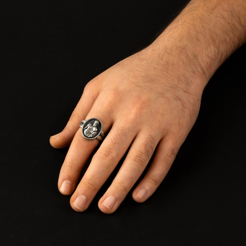 Puede incluir: Un anillo de plata con un camafeo ovalado negro que presenta una imagen detallada de una persona. El anillo se lleva en el dedo &iacute;ndice de una mano.