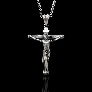 May include: A silver-colored crucifix pendant with a detailed depiction of Jesus Christ on the cross. The pendant hangs from a delicate silver chain. The background is solid black, highlighting the pendant's intricate design.