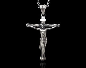 Può includere: Un ciondolo a forma di crocifisso color argento con una dettagliata raffigurazione di Gesù Cristo sulla croce. Il ciondolo è appeso a una delicata catena d'argento. Lo sfondo è nero, evidenziando il design intricato del ciondolo.