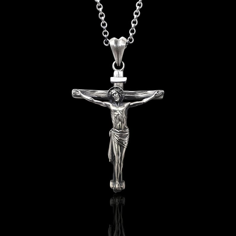 May include: A silver-colored crucifix pendant with a detailed depiction of Jesus Christ on the cross. The pendant hangs from a delicate silver chain. The background is solid black, highlighting the pendant's intricate design.