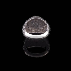 Op de afbeelding: Zilveren ring met een ingewikkeld ontwerp met een gevleugeld wezen en een inscriptie. De ring wordt van binnenuit getoond, waardoor de details van het ontwerp zichtbaar worden.