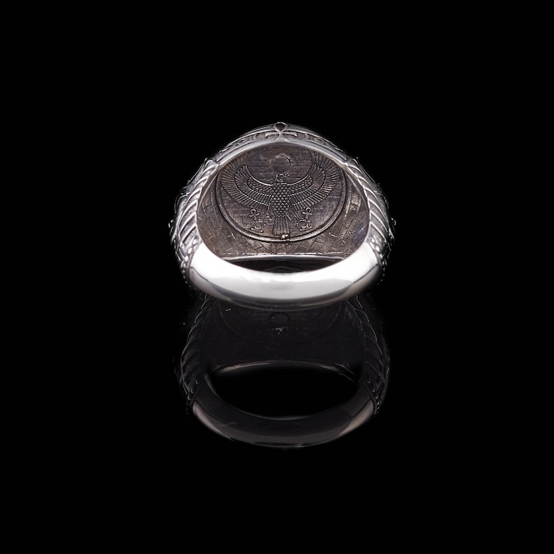 Op de afbeelding: Zilveren ring met een ingewikkeld ontwerp met een gevleugeld wezen en een inscriptie. De ring wordt van binnenuit getoond, waardoor de details van het ontwerp zichtbaar worden.