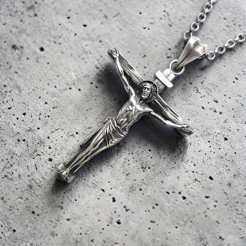 May include: A silver-colored crucifix pendant with a detailed depiction of Jesus Christ on the cross. The pendant hangs from a delicate chain with a heart-shaped clasp. The cross is made of a silver-toned metal.