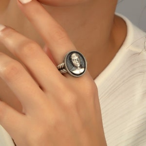 May include: A silver ring with a black oval cameo featuring a detailed portrait of a person. The ring has a rope-like band.