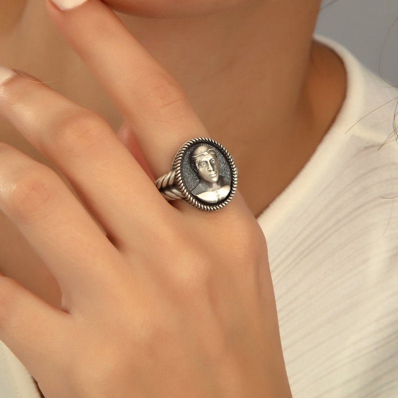 May include: A silver ring with a black oval cameo featuring a detailed portrait of a person. The ring has a rope-like band.