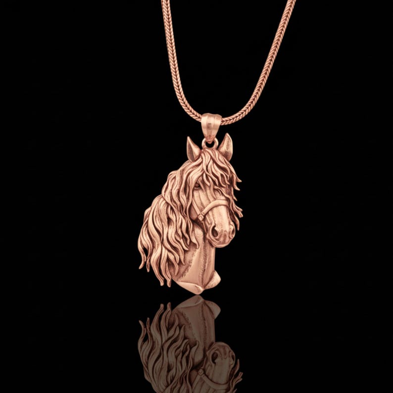 Puede incluir: Un collar con colgante de caballo en oro rosa. El colgante presenta una talla detallada de la cabeza de un caballo con melena y brida fluidas. El collar tiene una cadena delicada. El fondo es negro.