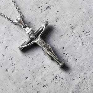 May include: A silver-colored crucifix pendant with a detailed depiction of Jesus Christ on the cross. The pendant hangs from a delicate chain. The background is a textured, gray surface.