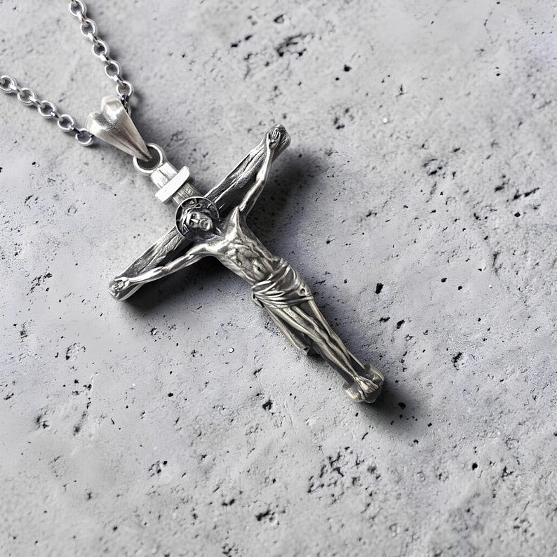 May include: A silver-colored crucifix pendant with a detailed depiction of Jesus Christ on the cross. The pendant hangs from a delicate chain. The background is a textured, gray surface.
