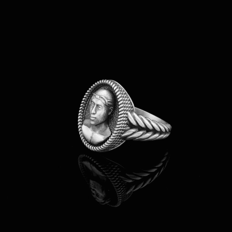 May include: A silver ring with a detailed design. The ring features a raised oval cameo with a sculpted image of a person's face.