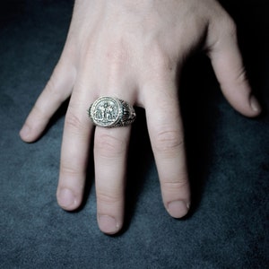 Op de afbeelding: Een zilveren ring met een ingewikkeld ontwerp met twee figuren die een weegschaal vasthouden. De ring wordt gedragen aan de middelvinger van een hand.