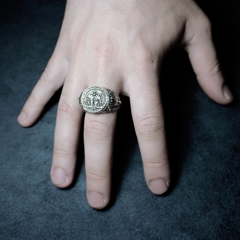 Op de afbeelding: Een zilveren ring met een ingewikkeld ontwerp met twee figuren die een weegschaal vasthouden. De ring wordt gedragen aan de middelvinger van een hand.