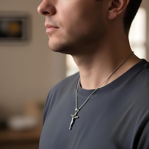 May include: Silver-toned crucifix pendant necklace. The pendant depicts a figure on a cross. The necklace is worn on a person wearing a gray t-shirt. The chain is a delicate, linked design.