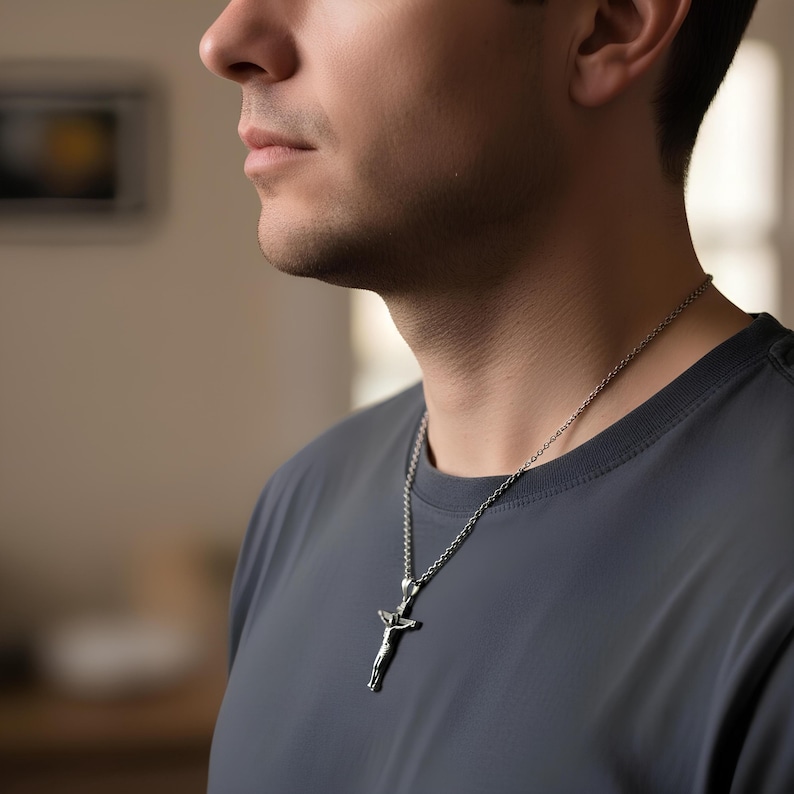 May include: Silver-toned crucifix pendant necklace. The pendant depicts a figure on a cross. The necklace is worn on a person wearing a gray t-shirt. The chain is a delicate, linked design.