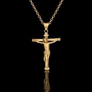 May include: A gold-colored pendant necklace featuring a crucifix with a detailed depiction of Jesus Christ. The pendant hangs from a delicate chain. The background is black, highlighting the jewelry.