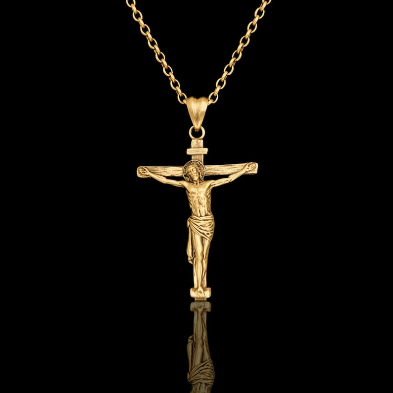 May include: A gold-colored pendant necklace featuring a crucifix with a detailed depiction of Jesus Christ. The pendant hangs from a delicate chain. The background is black, highlighting the jewelry.