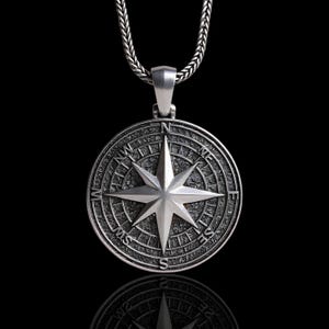 May include: A silver compass pendant necklace. The pendant showcases a detailed compass rose design with directional markers and an eight-pointed star. The necklace has a textured chain and a polished finish, set against a black backdrop.