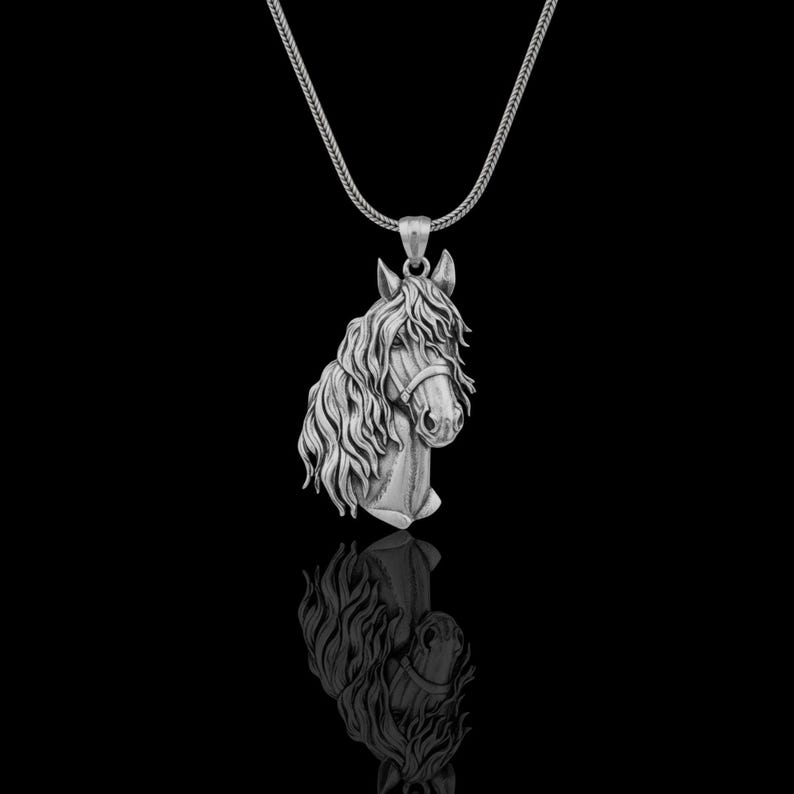 Puede incluir: Collar con colgante de plata con la cabeza de un caballo detallada. El caballo tiene una melena fluida y lleva un cabestro. El colgante cuelga de una delicada cadena sobre un fondo negro.