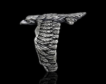 May include: A silver-toned ring shaped like a soaring eagle with wings extended horizontally. The metal surface features detailed, dark-oxidised engravings that define the individual feathers and the eagle's head, creating a textured appearance.