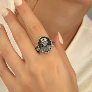 May include: A silver ring with a black and white portrait of a person wearing a hat. The ring has a rope-like design around the edge.
