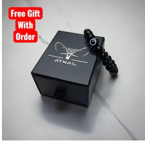 May include: A black bead bracelet with an evil eye charm rests on a black box with the AYNAL logo. The box has a small drawer and the words "Free Gift With Order" are in a red box in the upper left corner.