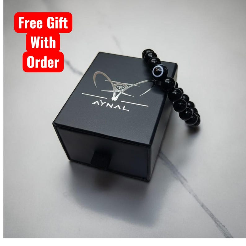 May include: A black bead bracelet with an evil eye charm rests on a black box with the AYNAL logo. The box has a small drawer and the words "Free Gift With Order" are in a red box in the upper left corner.