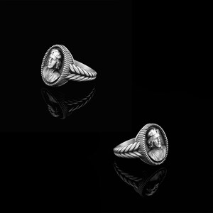 May include: Two silver rings with a detailed design of a person's face on the front. The rings are on a black background.