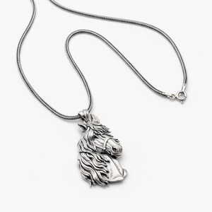 Puede incluir: Un collar con colgante plateado con un dise&ntilde;o detallado de cabeza de caballo. La cabeza del caballo est&aacute; esculpida con una melena fluida y detalles de brida. La cadena es una cadena texturizada tipo serpiente con cierre de broche. El colgante est&aacute; sobre un fondo blanco.