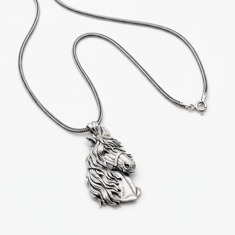 Puede incluir: Un collar con colgante plateado con un dise&ntilde;o detallado de cabeza de caballo. La cabeza del caballo est&aacute; esculpida con una melena fluida y detalles de brida. La cadena es una cadena texturizada tipo serpiente con cierre de broche. El colgante est&aacute; sobre un fondo blanco.