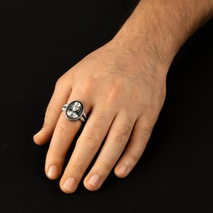 May include: A silver ring with a detailed oval-shaped design featuring a face. The ring is worn on a person's finger.