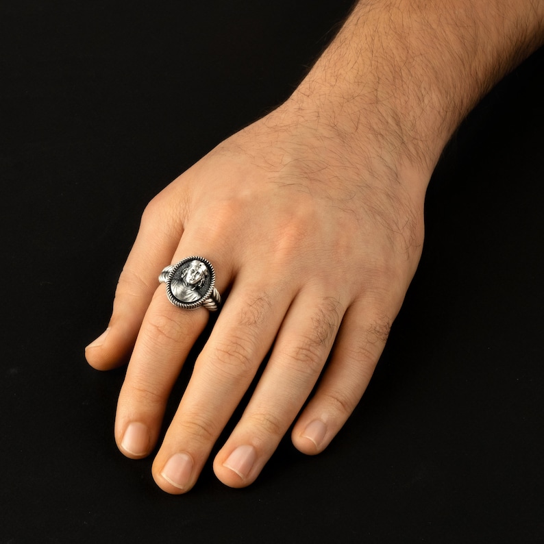 May include: A silver ring with a detailed oval-shaped design featuring a face. The ring is worn on a person's finger.