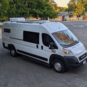 May include: A white van with a high roof and a white roof rack. The van has tinted windows and a black bumper.