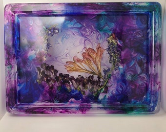 May include: Rectangular resin tray with a vibrant, swirled design in shades of purple, blue, and teal. The tray features dried flowers and a clear, glossy finish. The edges are rounded, and the overall aesthetic is artistic and decorative.