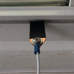 May include: Close-up of a grounding wire assembly. A black strap is secured to a white surface with a screw and copper connector. A white wire with a blue connector extends downward.