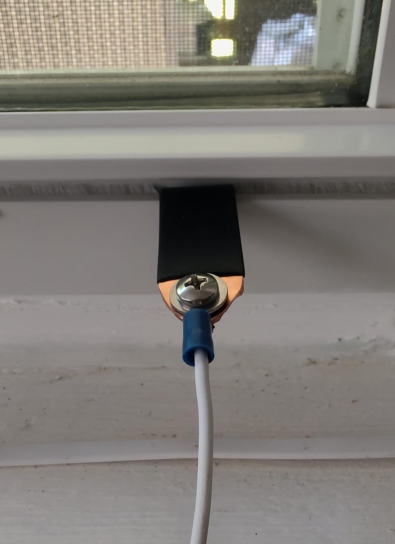 May include: Close-up of a grounding wire assembly. A black strap is secured to a white surface with a screw and copper connector. A white wire with a blue connector extends downward.
