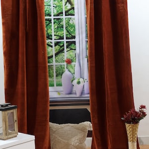May include: Two rust-coloured velvet curtains hang in a window with a white frame. The window has a view of a green tree and a white wall behind it. There is a white bedside table with two drawers and a white cushion on the floor.
