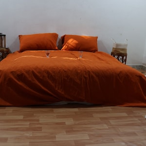 May include: A rust-colored linen duvet cover with two matching pillowcases on a bed. The bed is in a room with white walls and wooden floors.