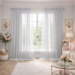 May include: A bright living room with pale blue, sheer curtains hanging from a gold curtain rail. The curtains frame a large window. The room includes a white rug, a sofa with pink cushions, and a glass-topped coffee table. Floral arrangements and framed art add decoration.