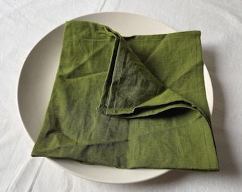 May include: A folded, olive green linen napkin rests on a white ceramic plate. The napkin is square, with visible texture and a slightly rumpled appearance. The plate is round with a wide rim, set against a white background.