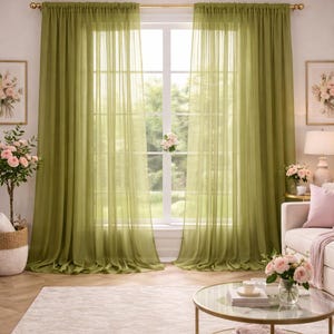 May include: Olive green sheer curtains hang in front of a window, allowing soft light to filter through. The curtains are suspended from a gold curtain rod. The room includes a light-coloured rug, a round coffee table, and a white sofa.