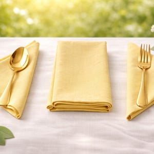 Puede incluir: Una mesa con tres servilletas amarillas y cubiertos dorados. Una cuchara descansa sobre una servilleta, un tenedor sobre otra, y la tercera servilleta está doblada. El fondo es una escena borrosa al aire libre.