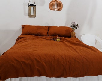 May include: A rust-coloured linen duvet cover with two matching pillowcases on a bed with a white bed skirt. The bed is in a bedroom with white walls and a white wicker chair.