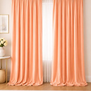 Op de afbeelding: Perzikkleurige fluwelen gordijnen die aan een gouden stang hangen. De gordijnen, die tot de vloer reiken, creëren een zachte, elegante uitstraling. De kamer heeft een neutraal kleurenpalet met een wit tapijt en een kleine bijzettafel.