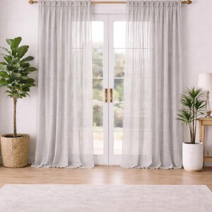 Puede incluir: Una habitación luminosa con cortinas grises claras y transparentes colgadas de una barra de madera. Una gran planta en maceta está a la izquierda y otra más pequeña a la derecha. Una alfombra gris clara cubre el suelo.