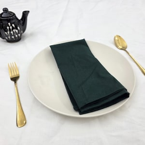 Pode incluir: Uma mesa posta com um prato branco, uma toalha de mesa verde-escura dobrada e talheres dourados. Uma pequena chaleira preta com detalhes brancos está ao lado.